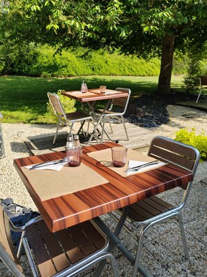 An outdoor wooden table with metal chairs, set on a gravel surface with a grassy area and trees in the background. - Campanile Caen Mondeville (Mondeville)