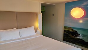 Habitación de hotel con una cama, una mesita de noche, una lámpara de pared y un cuadro de un atardecer sobre el océano.