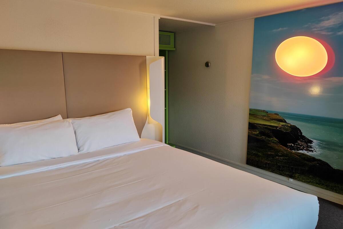 A hotel room with a bed, a nightstand, a wall-mounted lamp, and a painting of a sunset over the ocean.