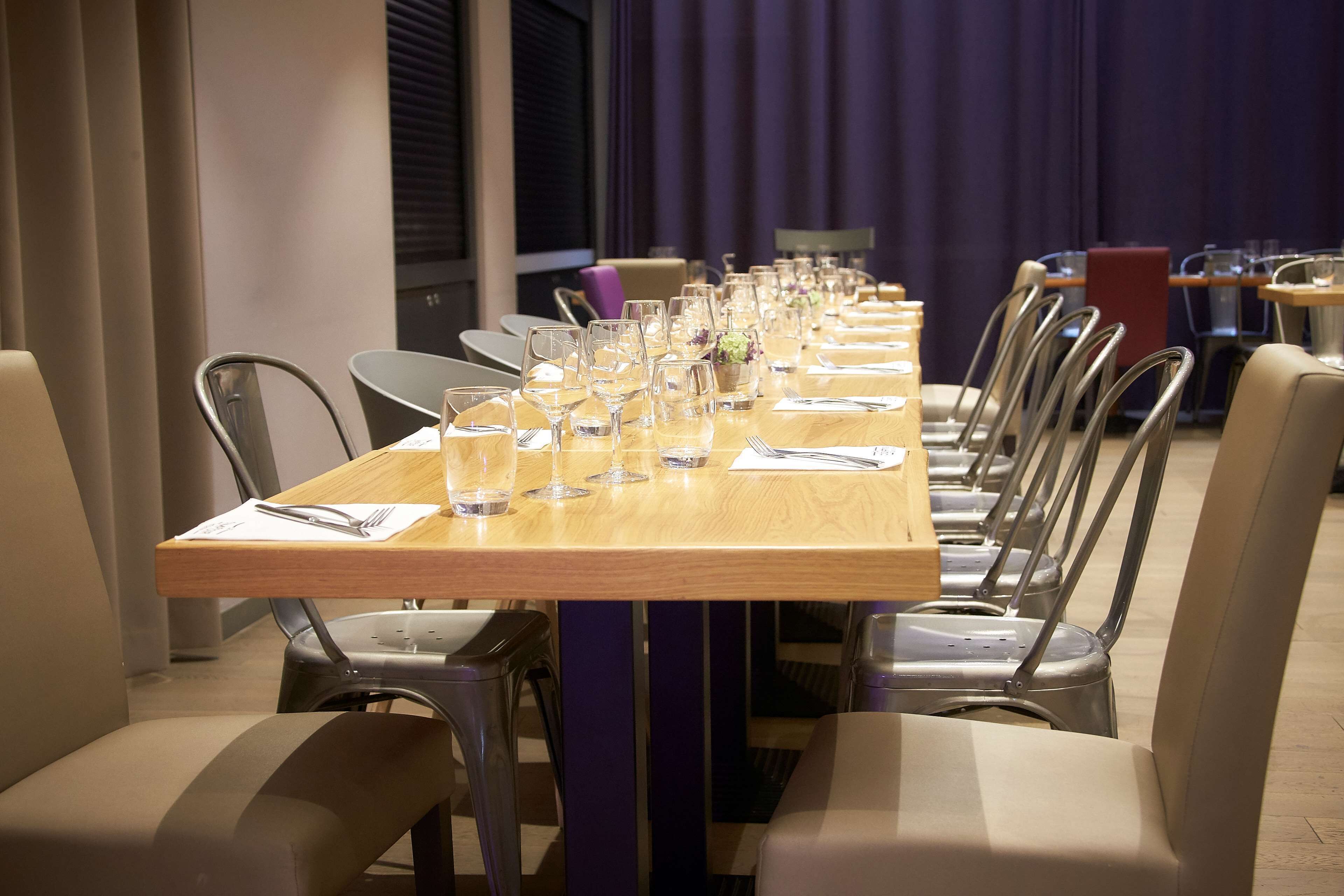 Una larga mesa de comedor con sillas, vasos y cubiertos, en una habitación con cortinas moradas y sillas modernas.