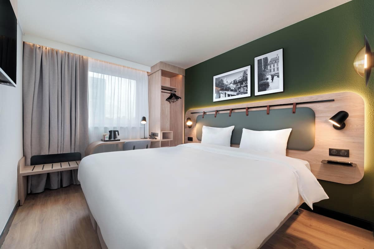 A modern hotel room with a large bed, a desk, a chair, a window with curtains, and two wall-mounted picture frames.