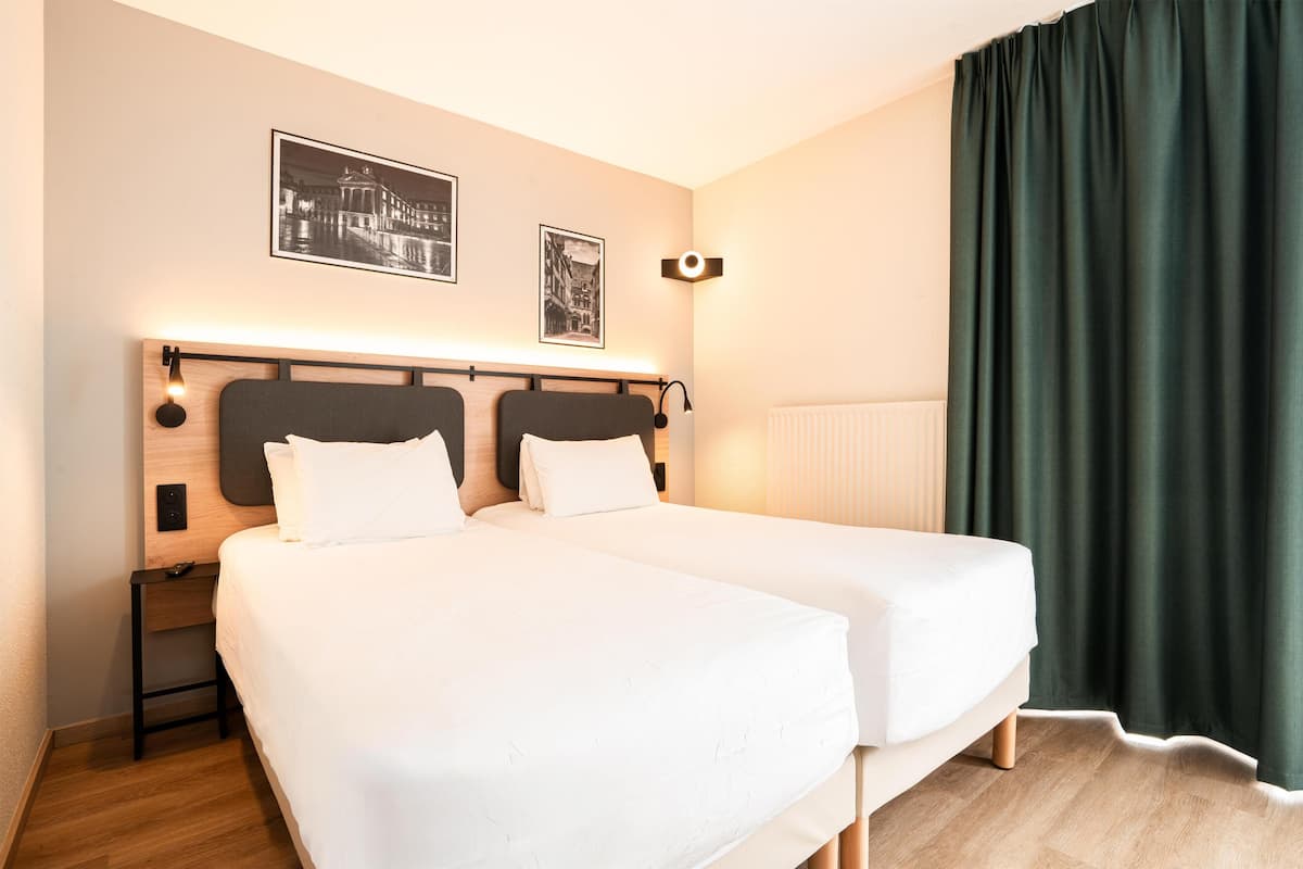 A hotel room with two beds, a nightstand, a wall-mounted lamp, and a window with curtains.