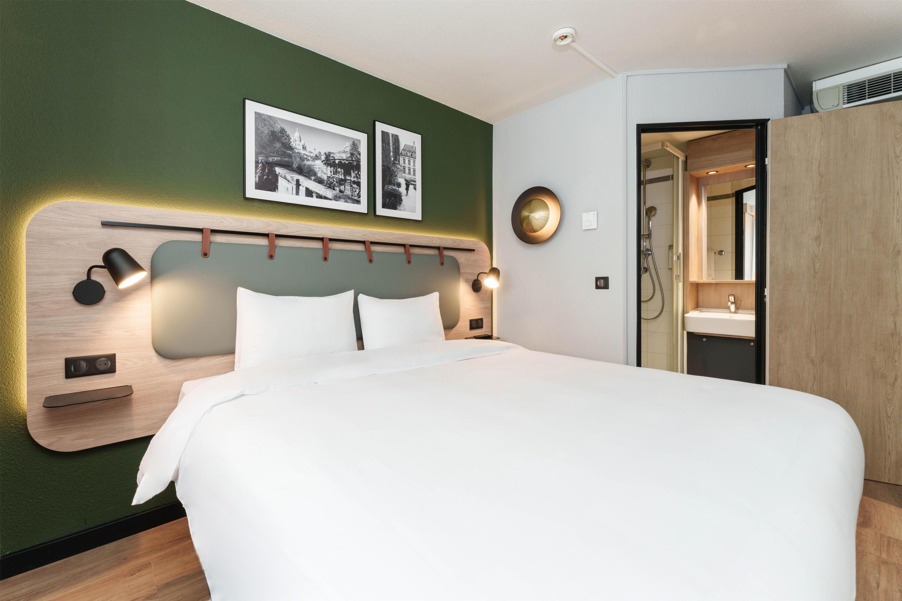 A modern hotel room with a large bed, a wooden headboard, wall-mounted lights, and a visible bathroom.