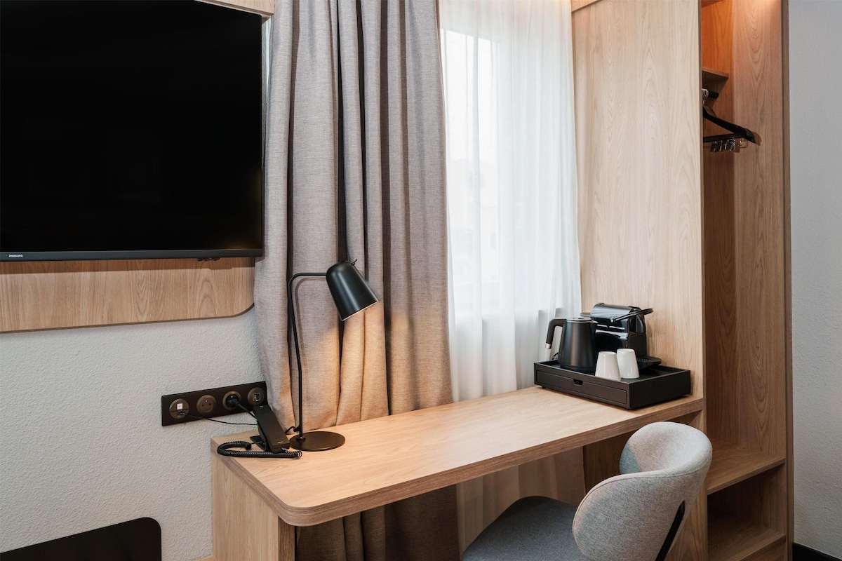 Quarto de hotel com escrivaninha de madeira, televisão de tela plana, luminária, cafeteira e uma cadeira.