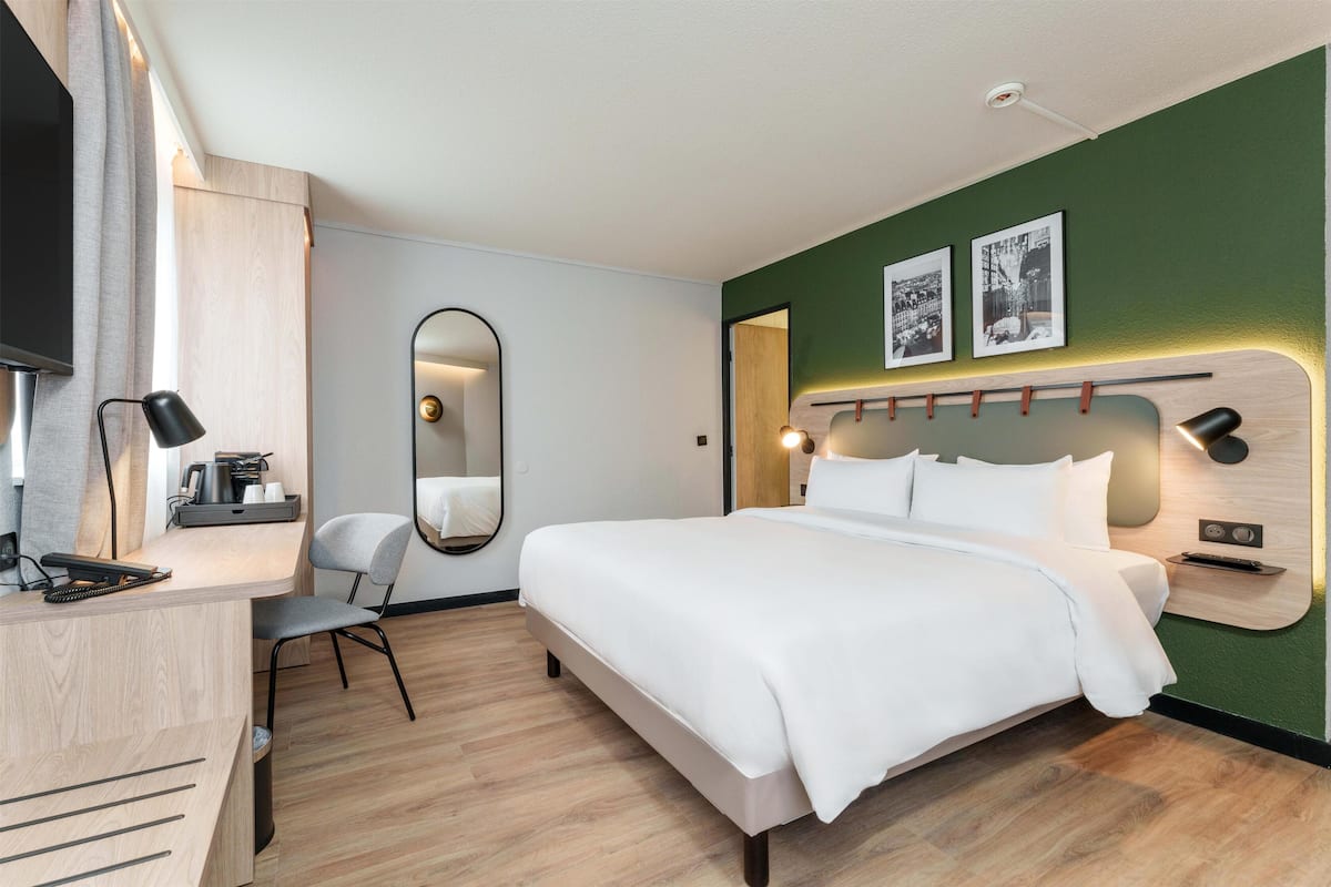 A modern hotel room with a large bed, a desk with a chair, a mirror, and a wall-mounted TV.