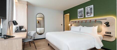 A modern hotel room with a large bed, a desk with a chair, a mirror, and a wall-mounted TV.