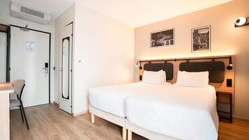 Une chambre d’hôtel avec un lit, un bureau et une chaise. Il y a une porte avec un miroir et un interrupteur. La chambre a un sol et des murs de couleur claire.