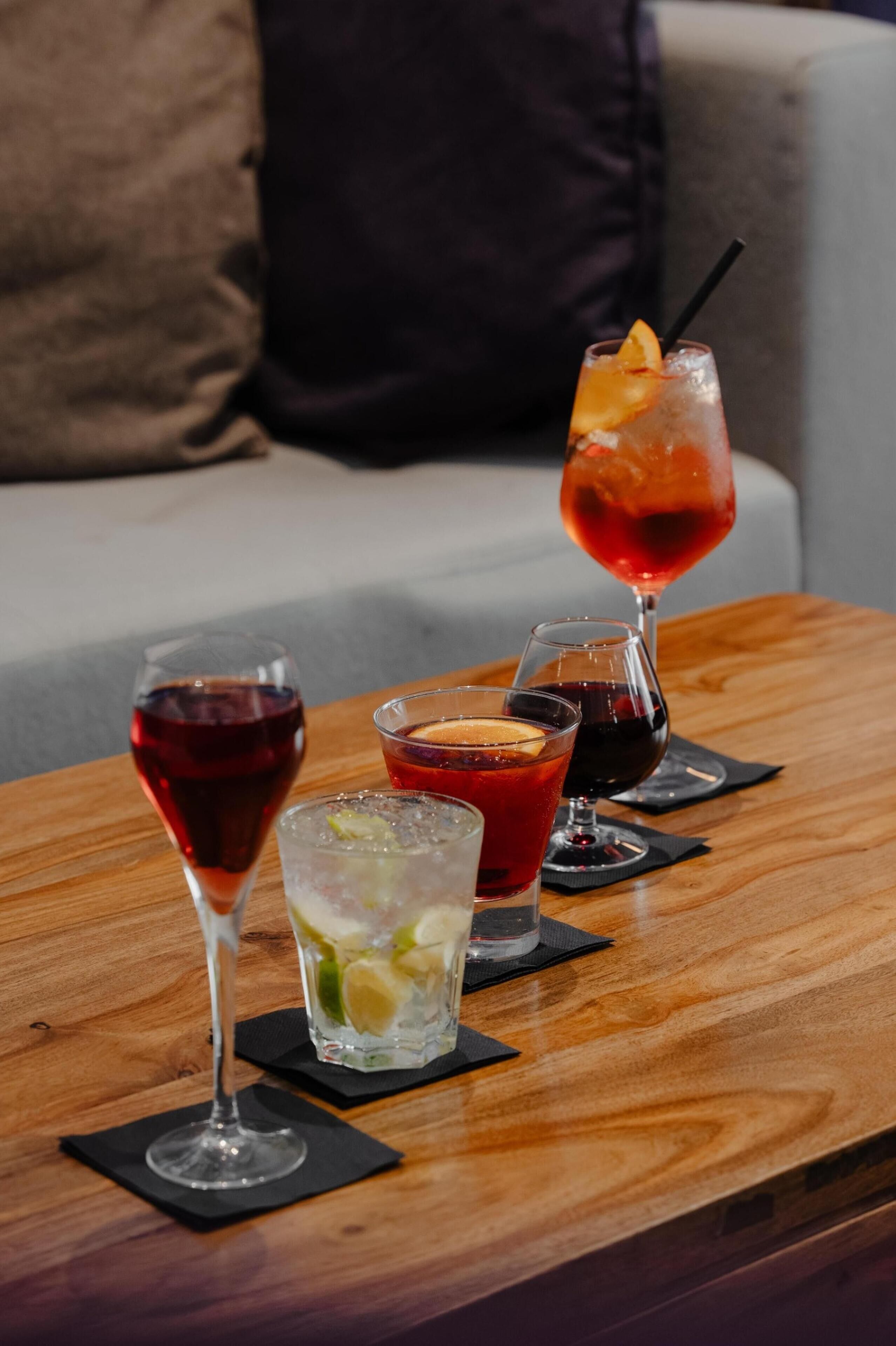 A wooden table with four glasses of various beverages, a black napkin, and a couch in the background.
