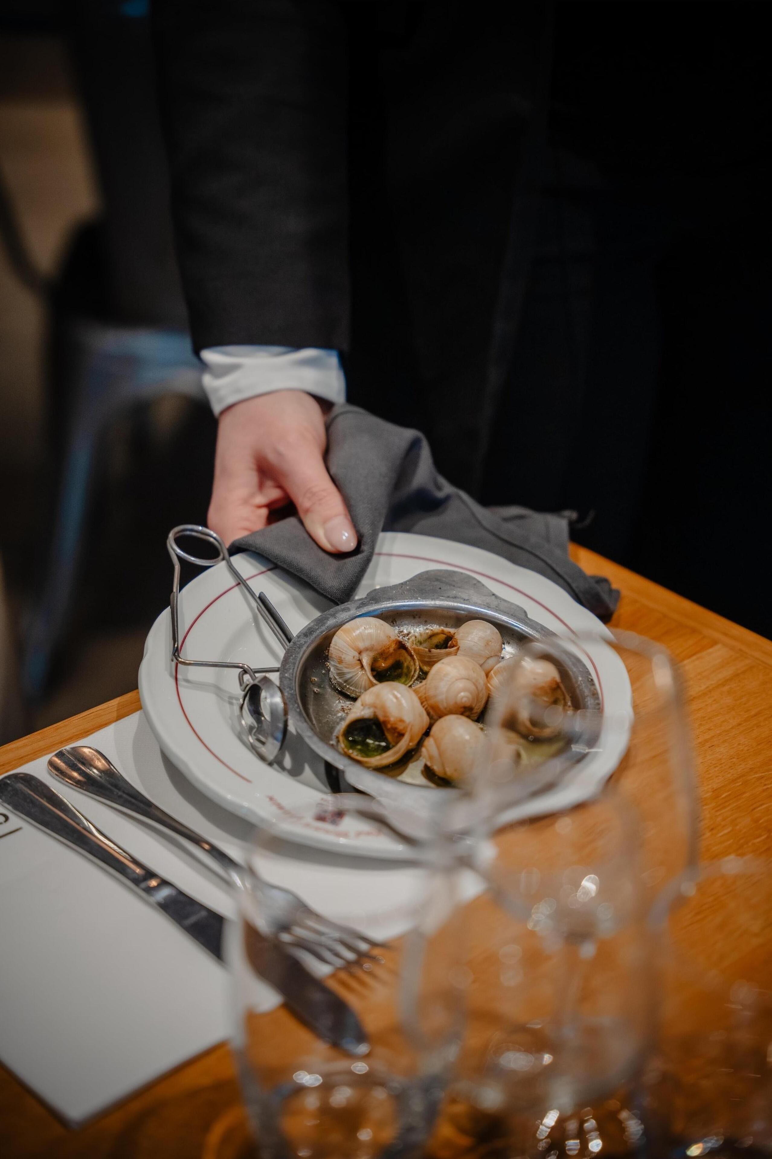 A dining table with a plate of escargot, a fork, a spoon, and a glass.