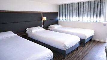 A hotel room with three single beds, a dark headboard, a bedside lamp, and a window with vertical blinds.