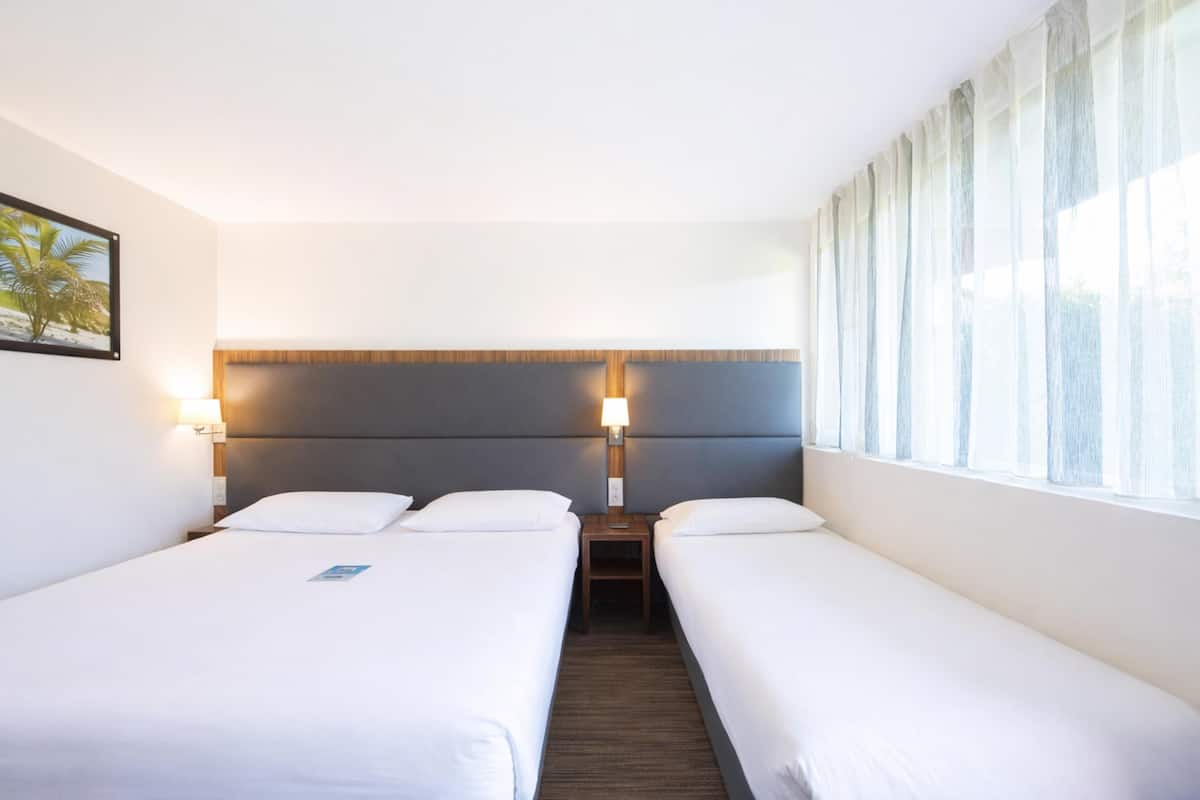 Une chambre d’hôtel avec deux lits simples, une table de chevet avec une lampe, une tête de lit et une fenêtre avec des rideaux.
