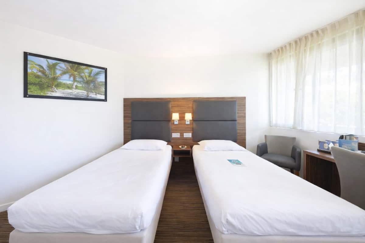 Une chambre d’hôtel avec deux lits simples, un bureau avec une chaise, une table de chevet, une grande fenêtre avec des rideaux et un tableau encadré au mur.