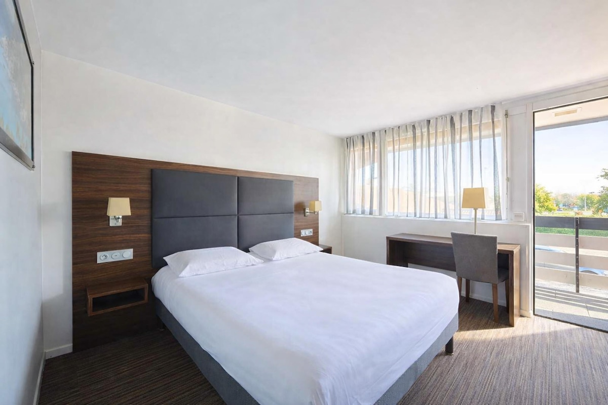 A hotel room with a large bed, a desk, a chair, and a window with curtains.