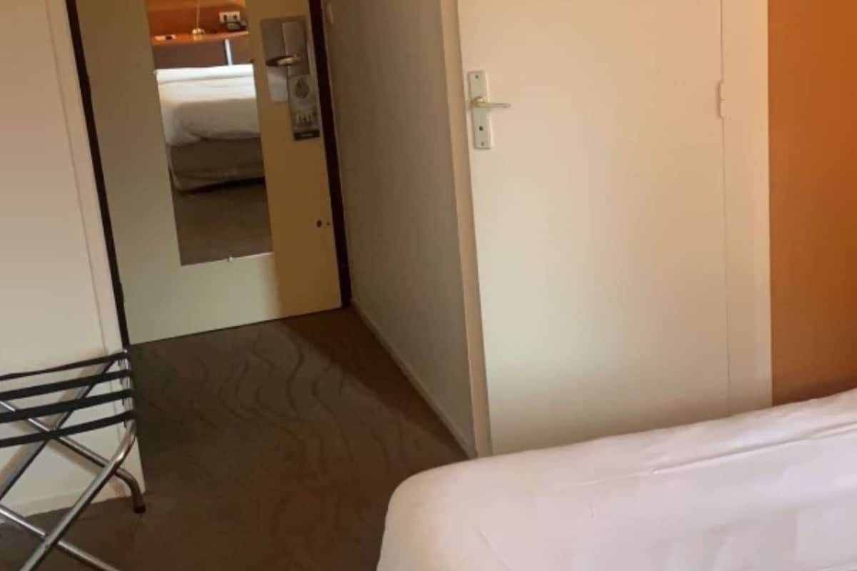 Une chambre d’hôtel avec un lit simple, une porte et une salle de bain visible, équipée d’un miroir et d’un lavabo.