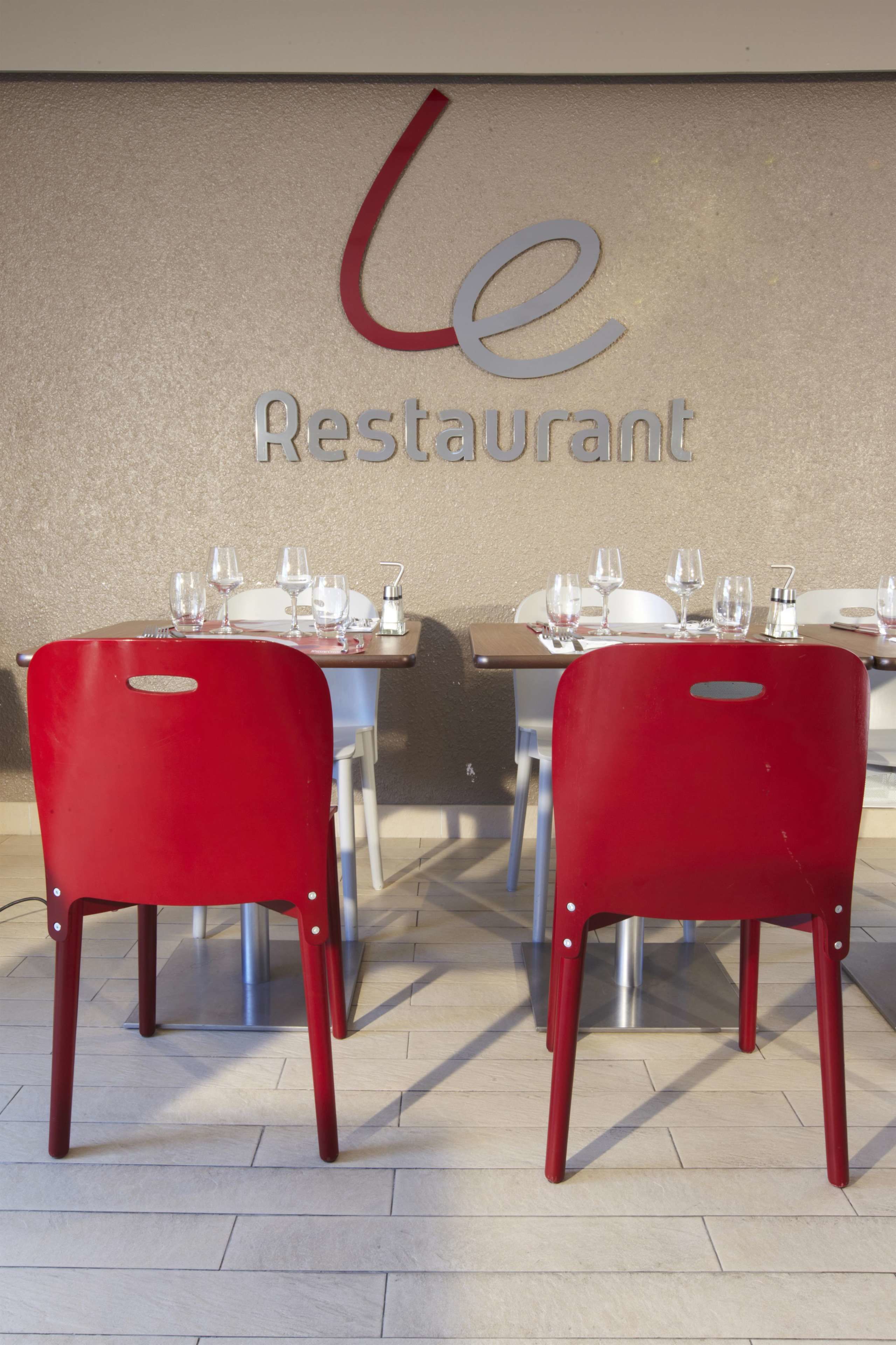 L’intérieur d’un restaurant, avec des chaises rouges, des tables dressées avec des verres à vin et des couverts, et une enseigne au mur indiquant « Le Restaurant ».