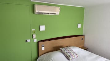 Une chambre d’hôtel avec un mur vert, une tête de lit en bois, un lit blanc avec un oreiller à motifs et un climatiseur fixé au mur.