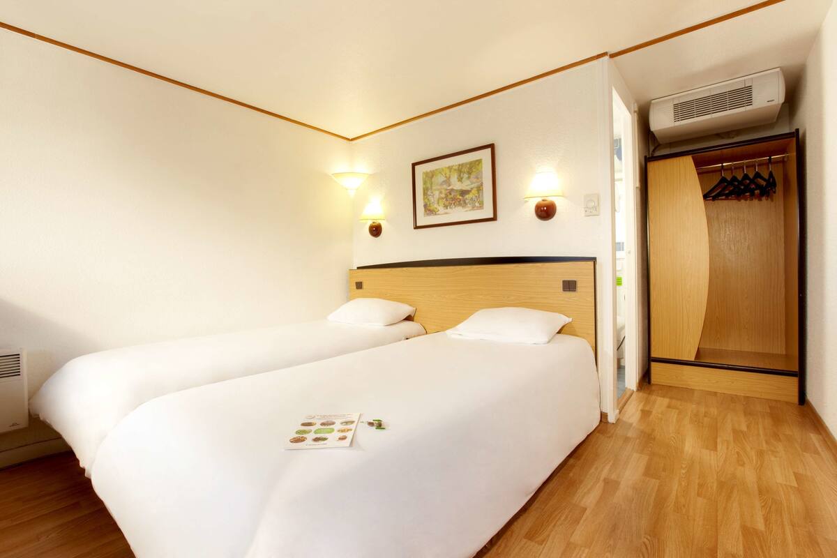 A hotel room with two beds, a wooden wardrobe, and a wall-mounted air conditioner.