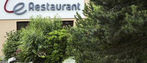 Un restaurant dont l’enseigne porte l’inscription « Le Restaurant », et qui est entouré de verdure et d’arbres.