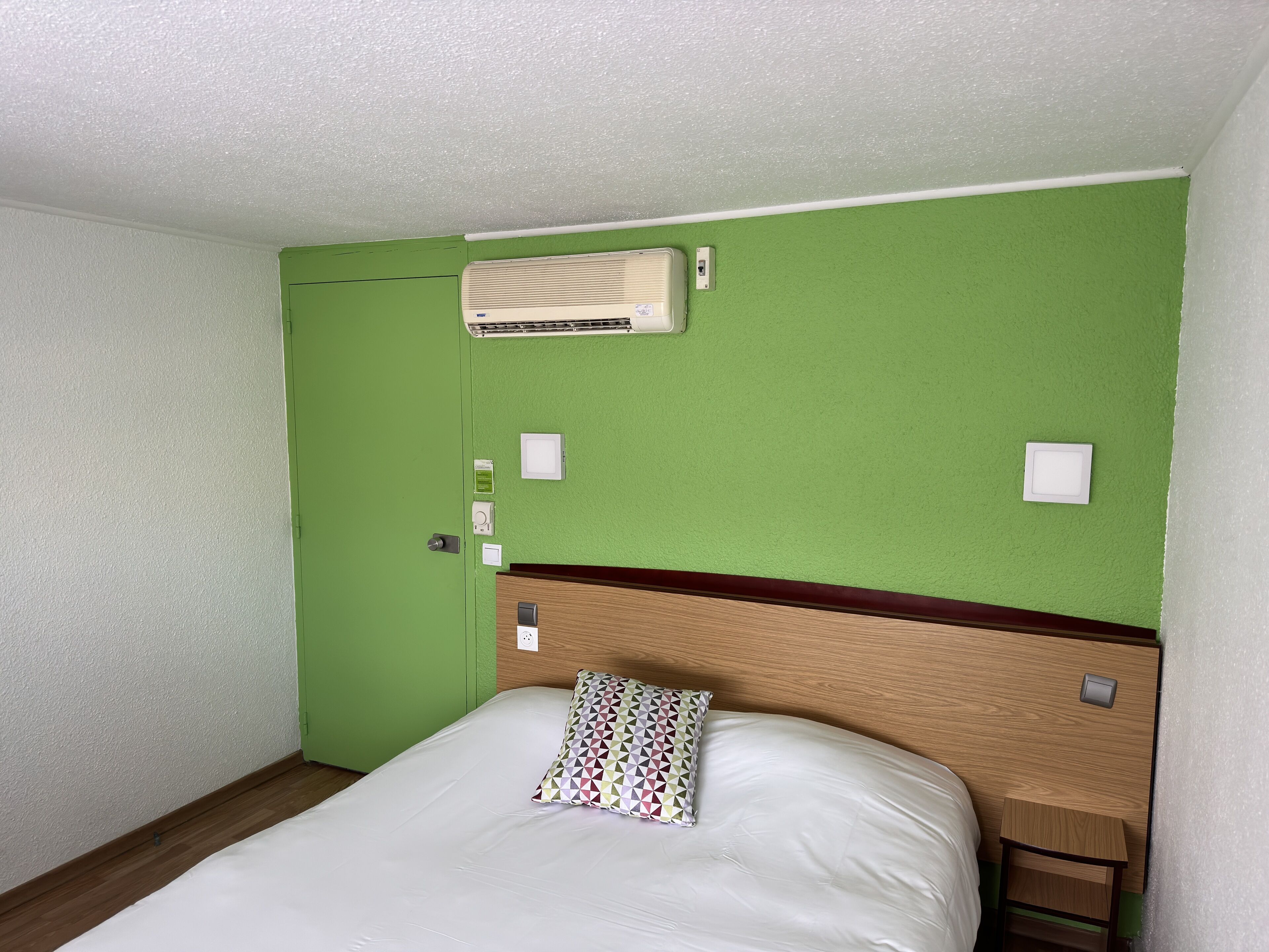 Habitación de hotel con pared verde, cabecero de madera, cama blanca, mesita de noche y aire acondicionado de pared.