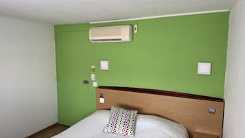 Habitación de hotel con pared verde, cabecero de madera, cama blanca, mesita de noche y aire acondicionado de pared.