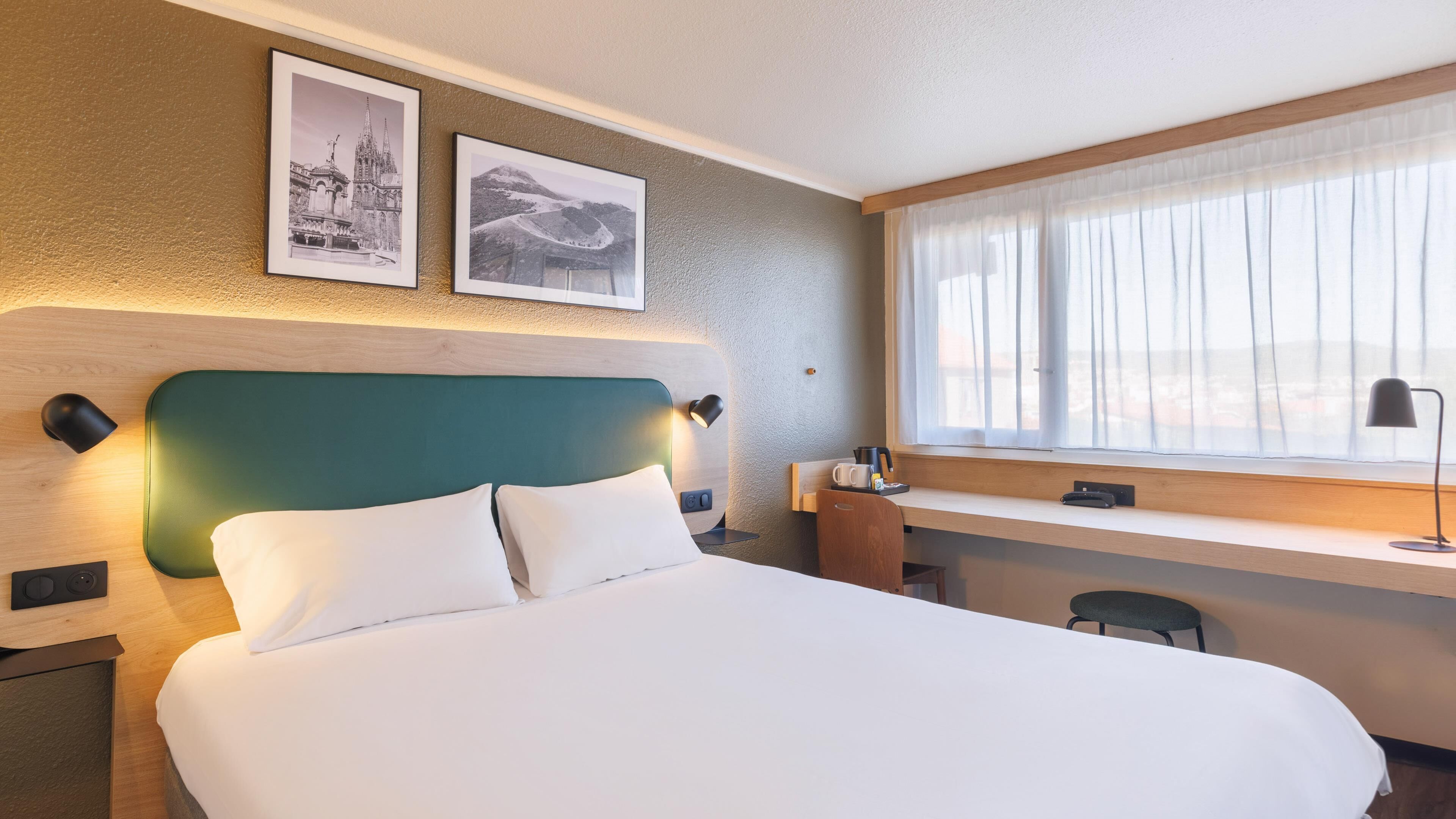 A hotel room with a bed, a desk, a chair, a lamp, and two framed pictures on the wall.