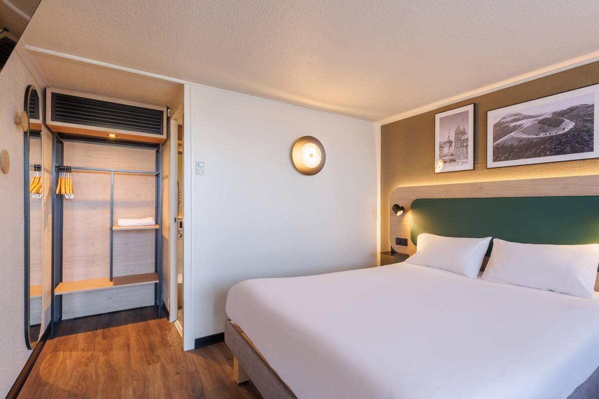 A hotel room with a bed, a wardrobe, a wall-mounted light, and two framed pictures.