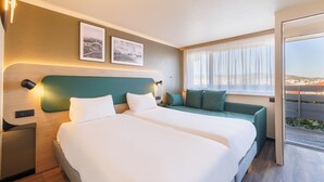 A hotel room with two beds, a sofa, a window with a view, and two framed pictures on the wall. - Campanile NATURE - Clermont Ferrand Le Brézet (Clermont-Ferrand)