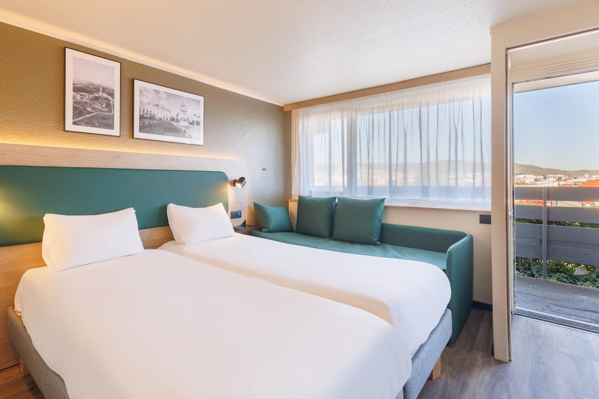 A hotel room with two beds, a sofa, a window with a view, and two framed pictures on the wall.