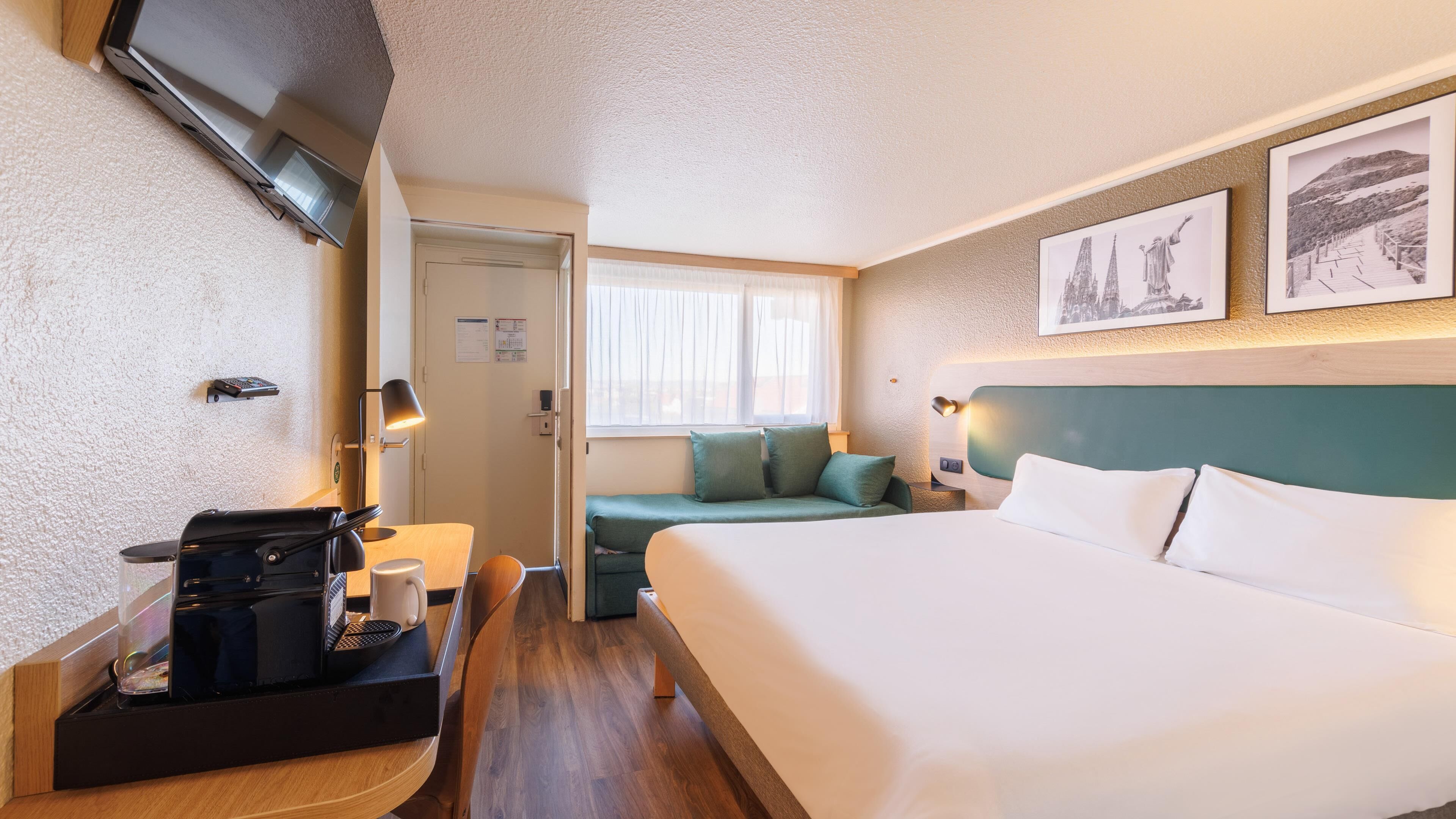 Una habitación de hotel con cama, escritorio con cafetera, zona de estar con cojines y cuadros en la pared.