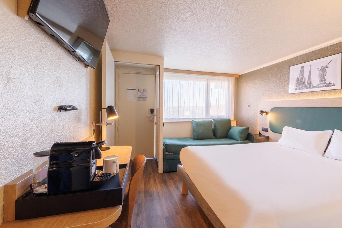 Ein Hotelzimmer mit Bett, Schreibtisch mit Kaffeemaschine, Sitzecke mit Kissen und gerahmten Bildern an der Wand.