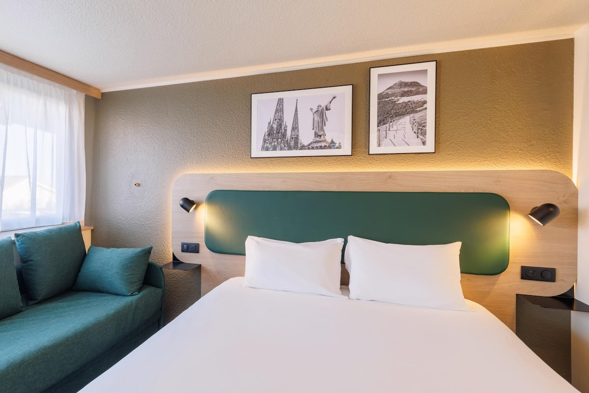 A hotel room with a bed, a green headboard, a teal sofa, two wall-mounted lamps, and two framed pictures.