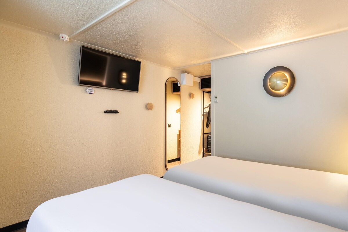 Ein Hotelzimmer mit einem Bett, einem an der Wand montierten Fernseher, einer runden Lampenleuchte und einem Kleiderhaken.