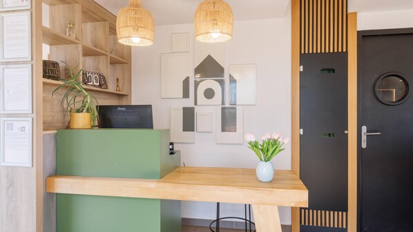 A reception desk with a green counter, a black device on it, and a vase with flowers. There are framed pictures on the wall and a black door with a round window.