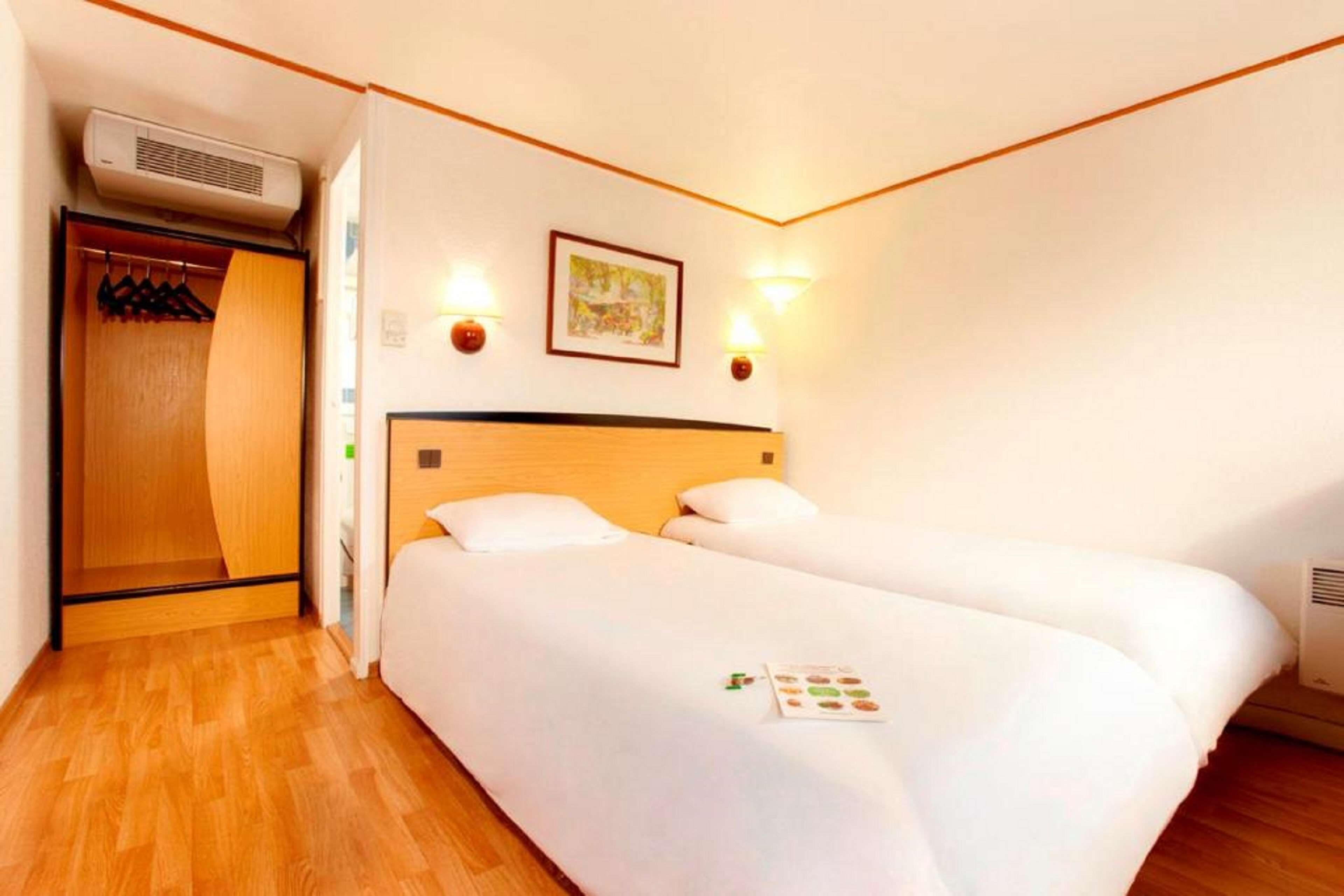 A hotel room with two single beds, a wooden wardrobe, a wall-mounted air conditioner, and two wall sconces.