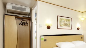 Una habitación de hotel con un armario de madera, una cama con ropa de cama blanca, una mesita de noche con un control remoto y dos lámparas fijadas a la pared.