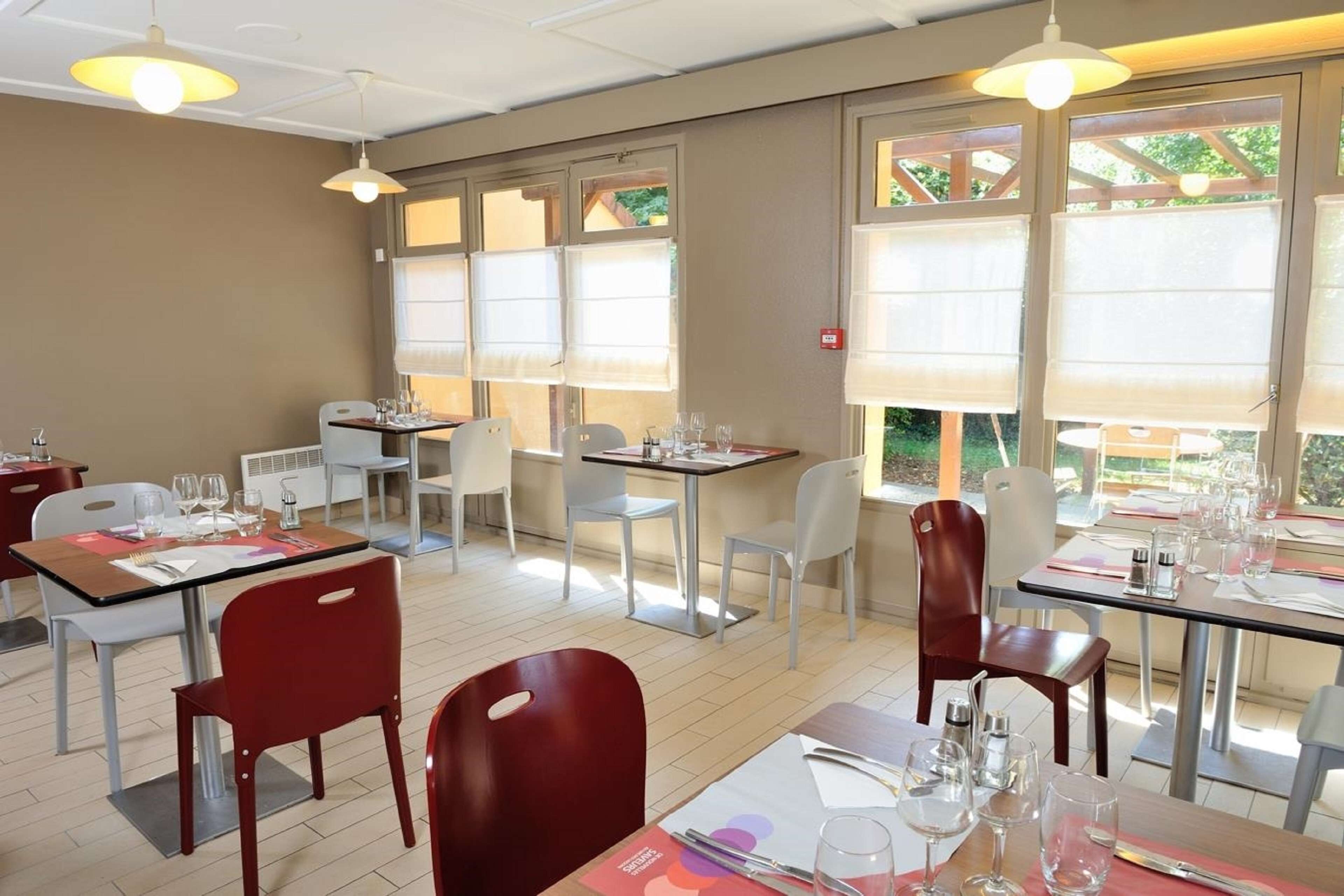 En restaurant med flere borde og stole, store vinduer med persienner og hængende lamper.