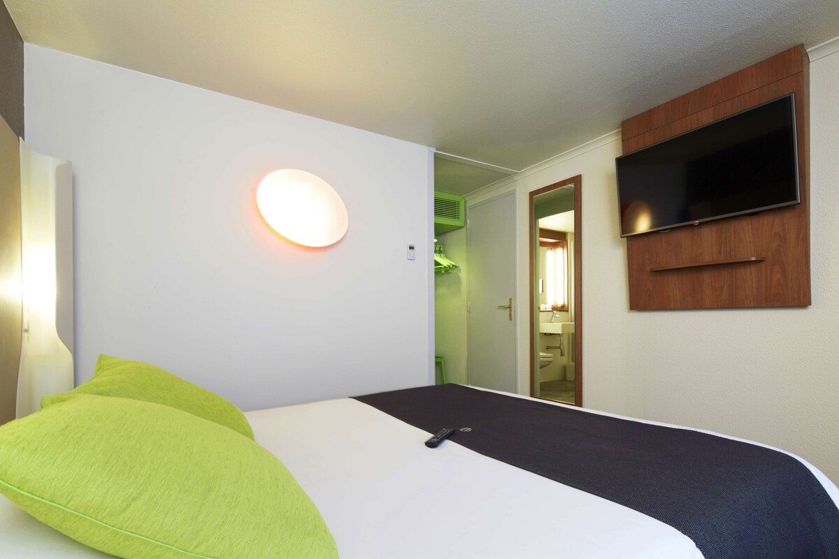 A hotel room with a bed, a TV mounted on the wall, and a round wall light.