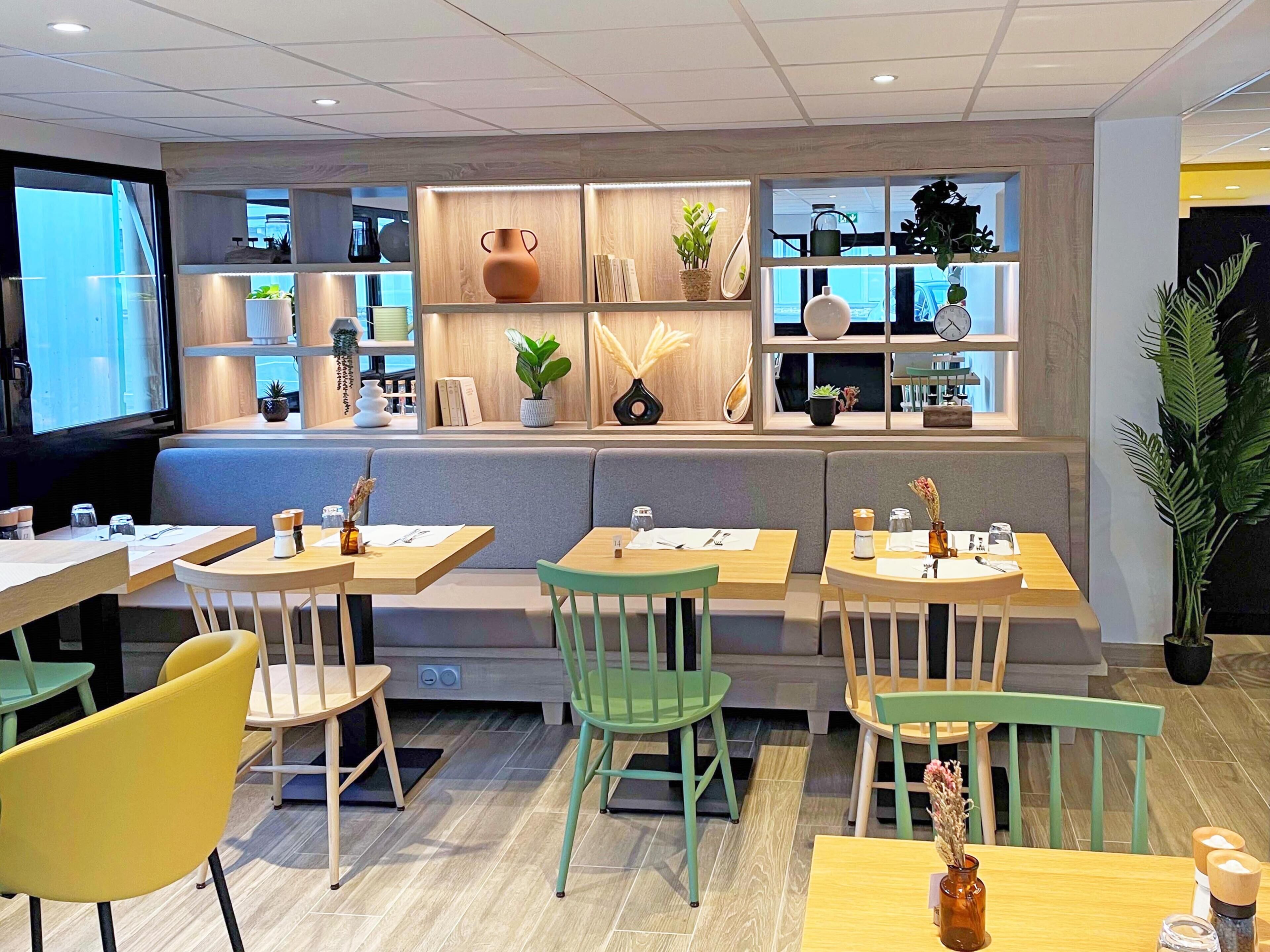 A modern restaurant with wooden furniture, green chairs, and a variety of plants on shelves.