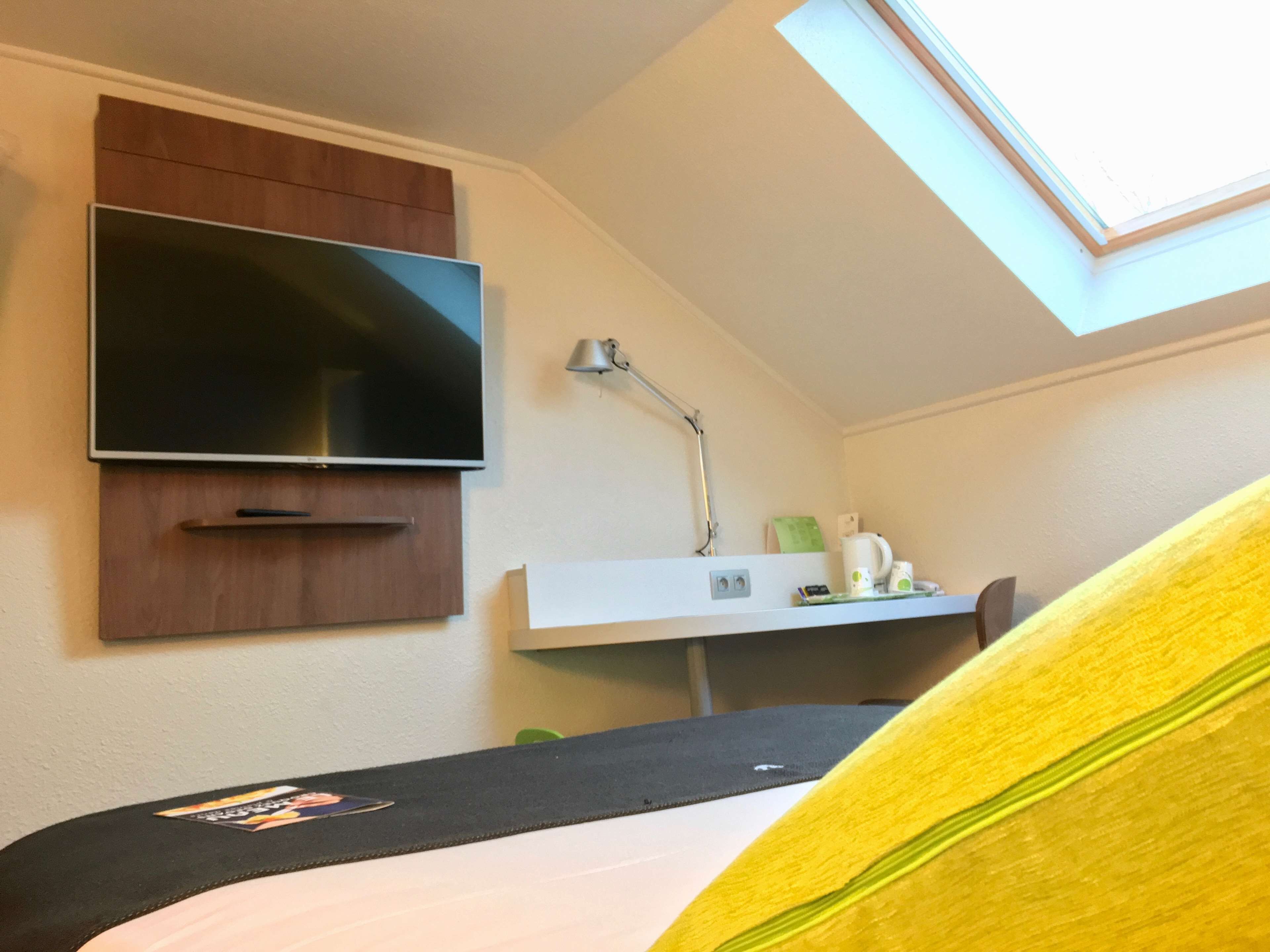 Ein Zimmer mit einem Bett, einem Schreibtisch, einem Flachbildfernseher an einer Holzvertäfelung, einer Schreibtischlampe und einem Oberlicht.