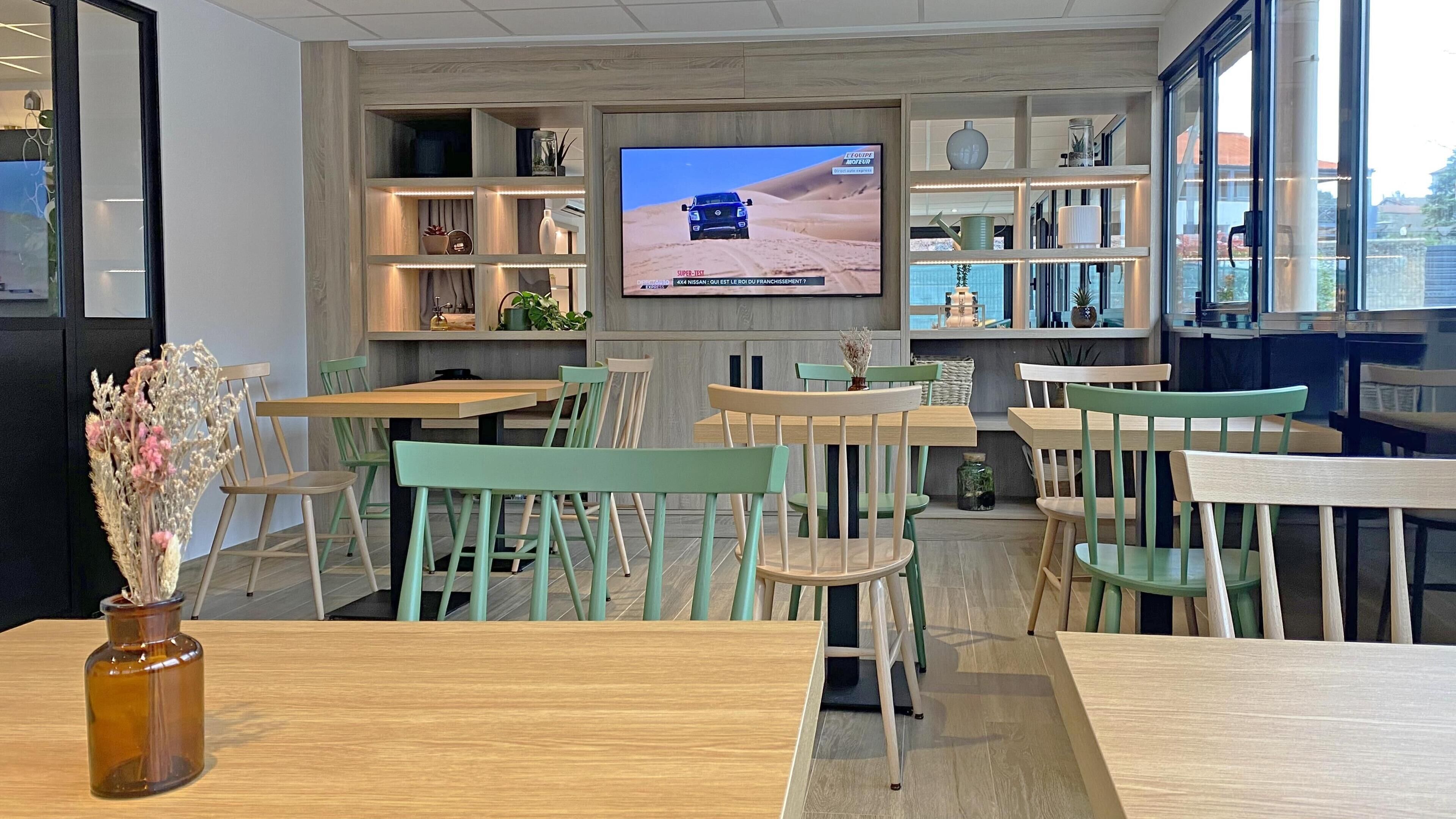 A modern interior with wooden tables and chairs, a television displaying a desert scene, and shelves with various items.