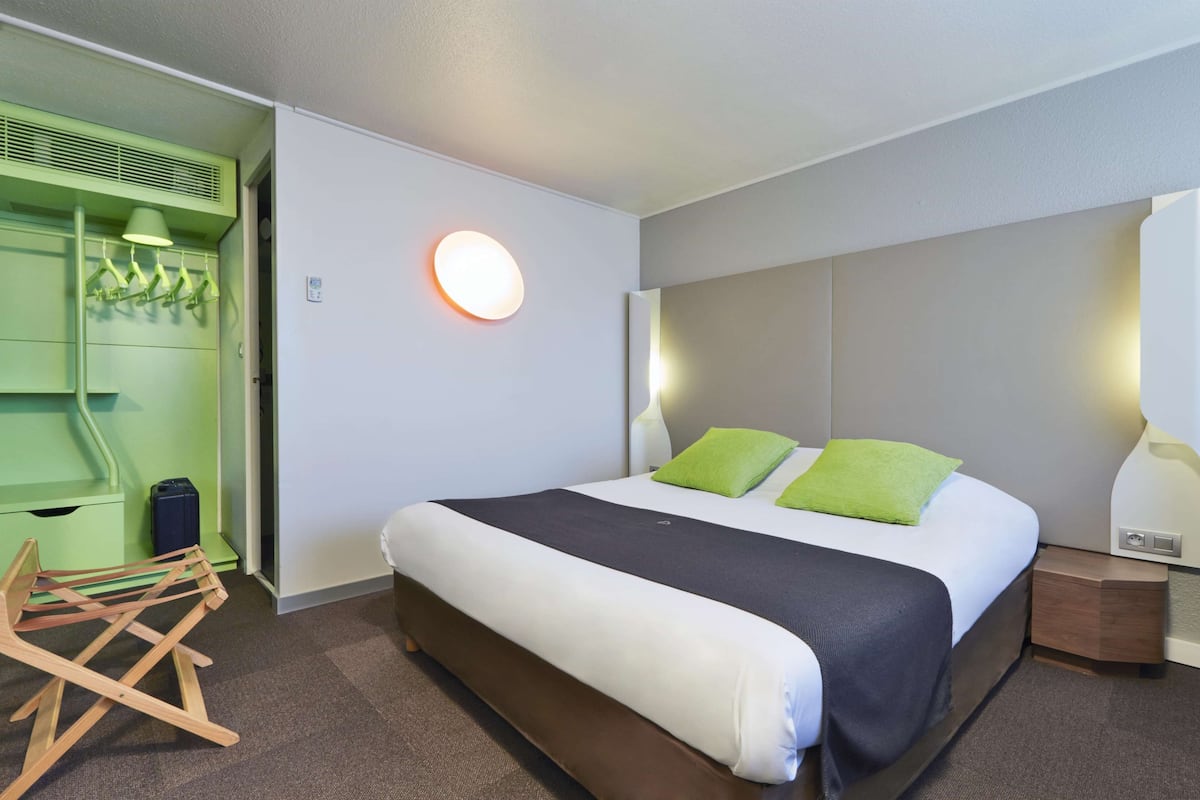 A modern hotel room with a neatly made bed, a green wall unit with hanging clothes, and a round wall light.