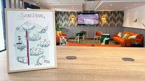 Un vestíbulo de hotel con una gran ilustración enmarcadas de Saint-Étienne, una zona de estar moderna con sofás naranja, una pantalla de televisión y un mostrador de recepción de madera.