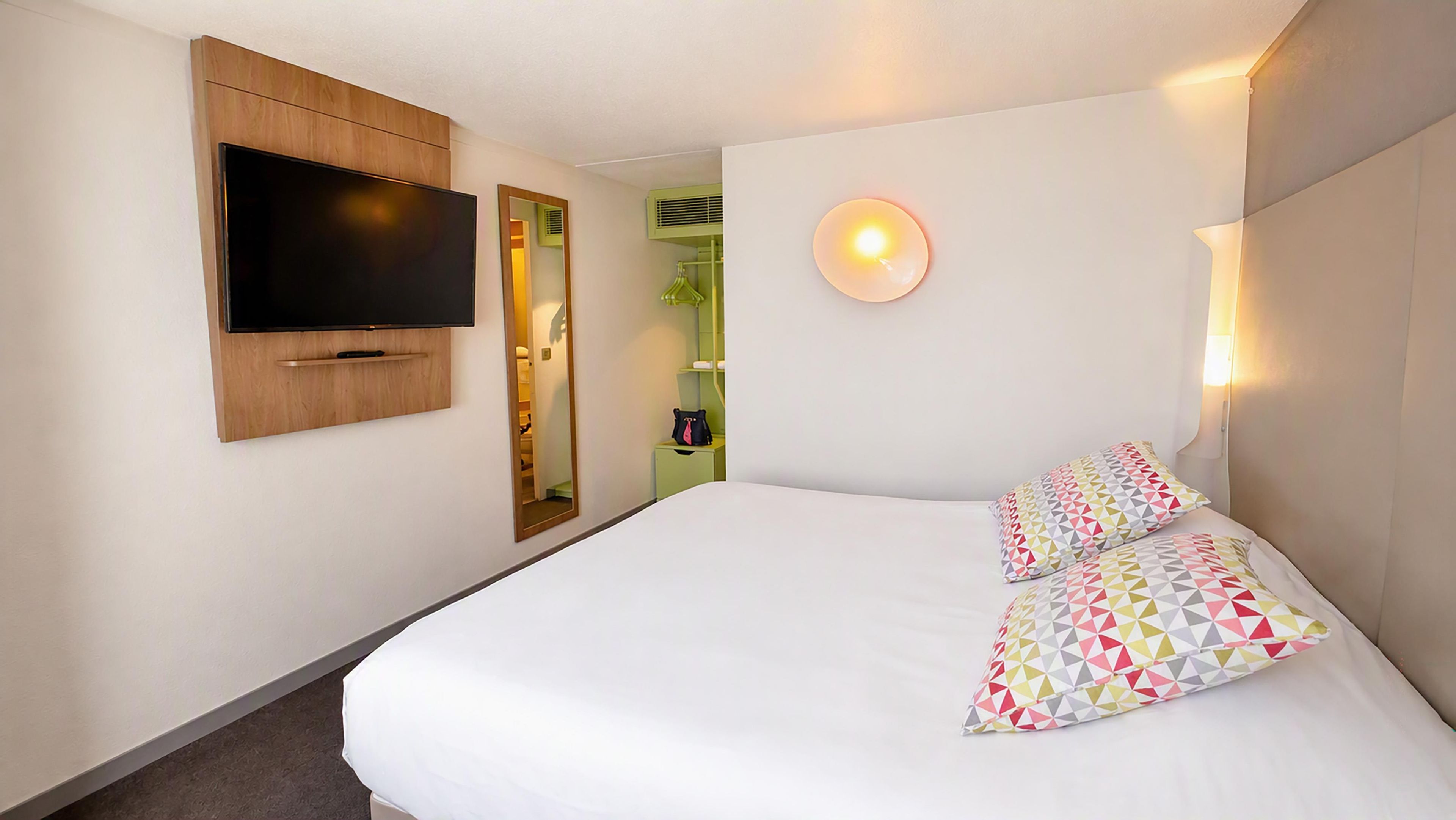 Habitación de hotel con cama, televisión montada en la pared, espejo y lámpara de noche.