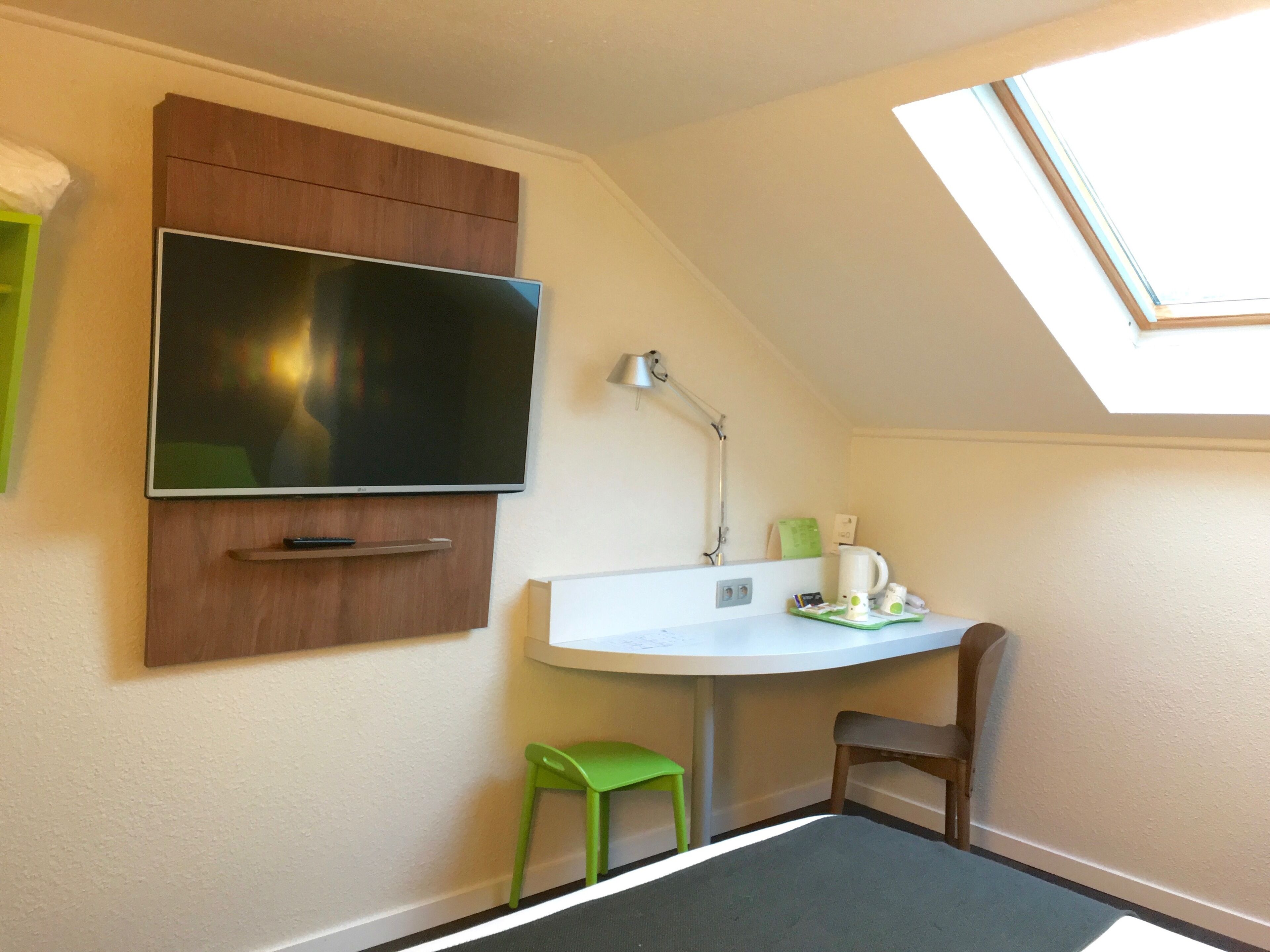 A small room with a TV mounted on the wall, a desk with a lamp, a chair, and a small stool.
