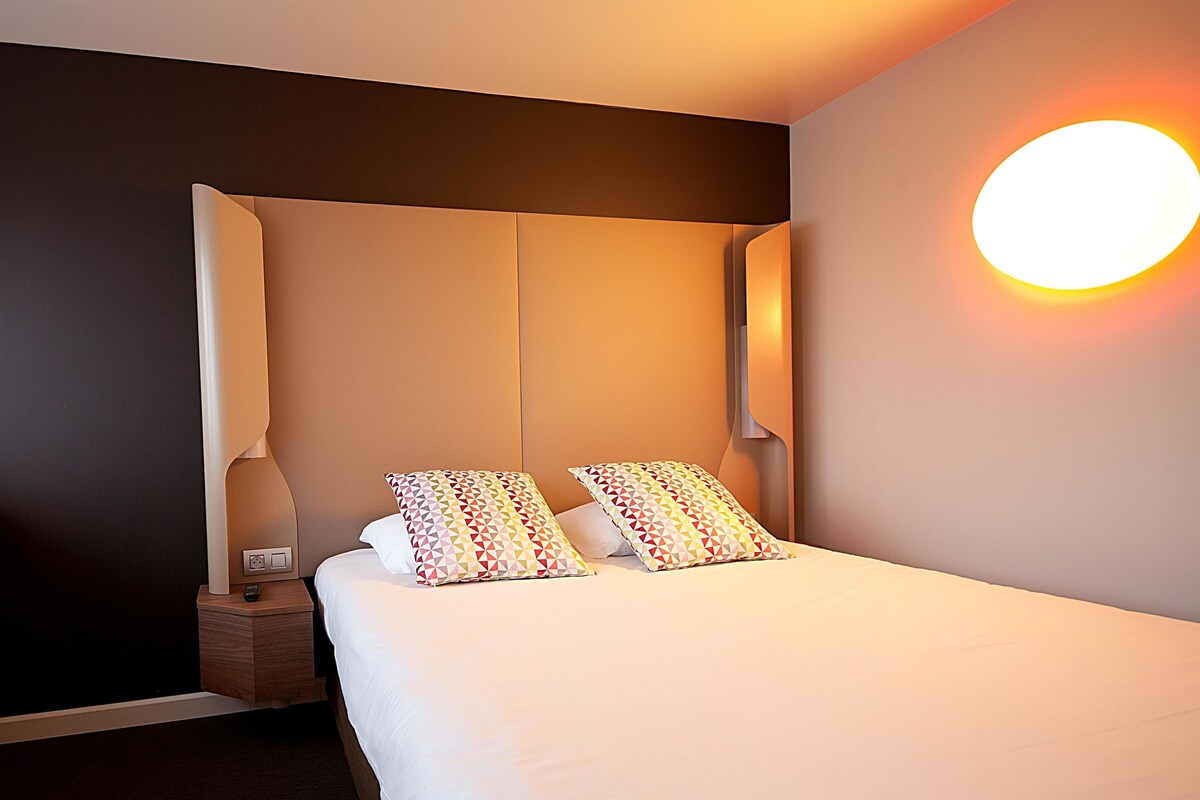 Une chambre d’hôtel moderne avec un grand lit, deux coussins à motifs, une table de chevet avec une lampe et un luminaire fixé au mur.