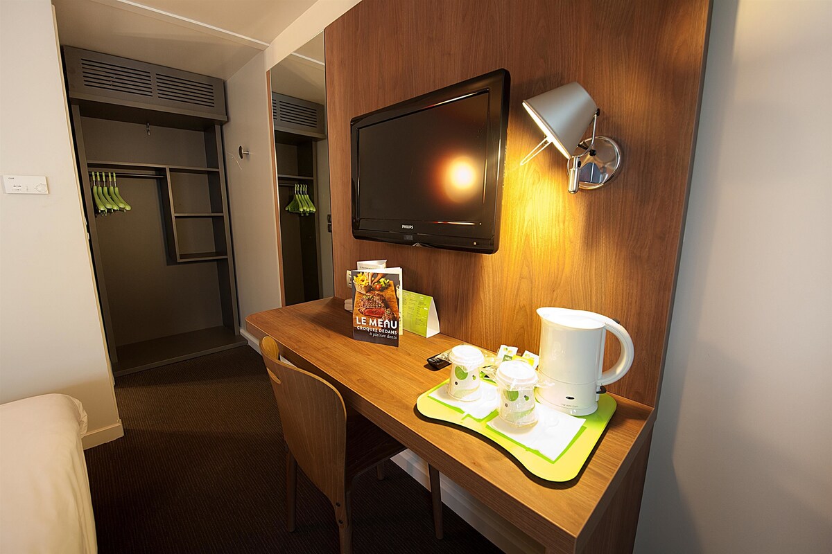 Une chambre d’hôtel avec un bureau en bois, un téléviseur à écran plat, une lampe fixée au mur, une machine à café et un menu posé sur la table.