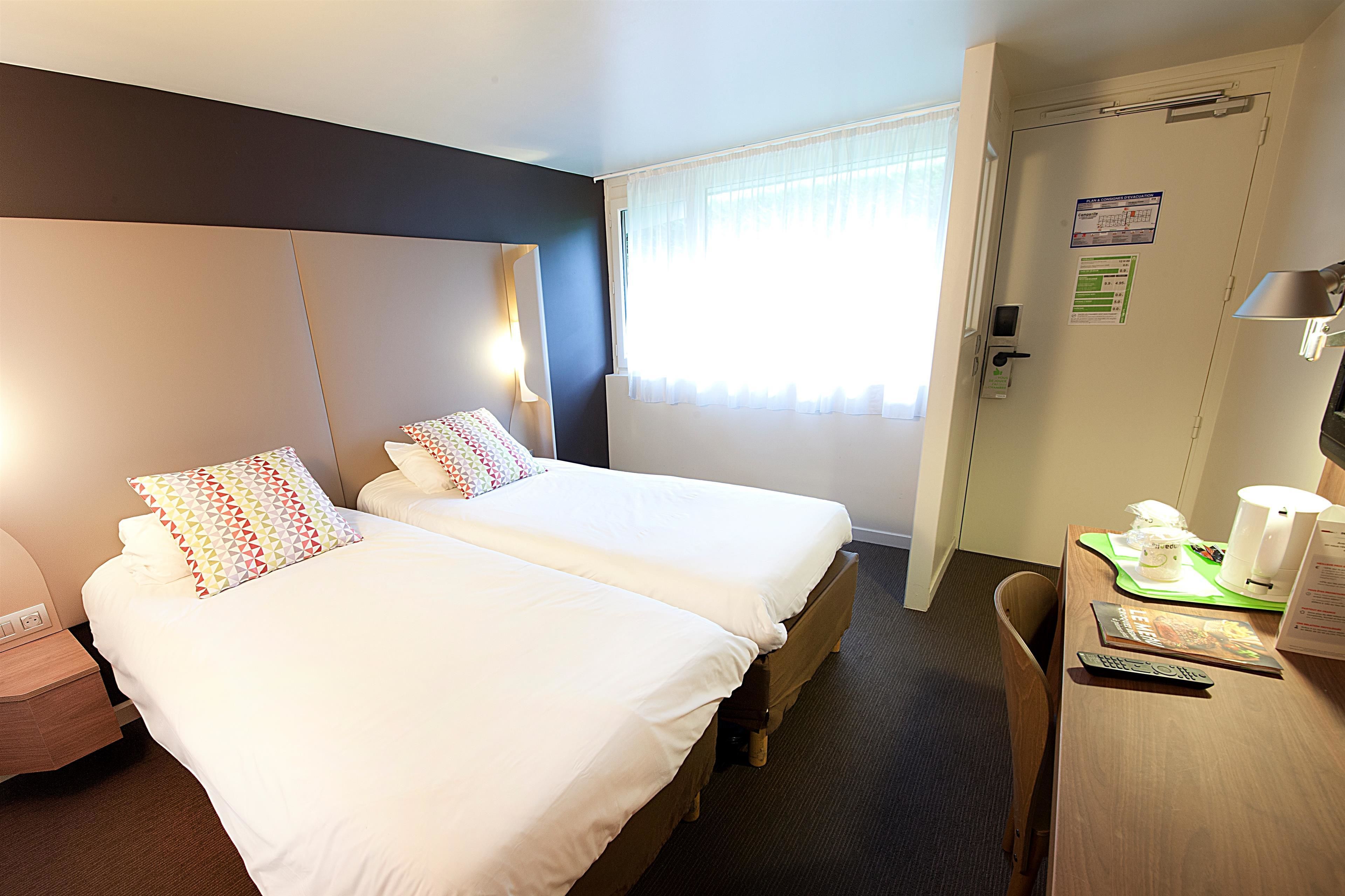 Quarto de hotel com duas camas, uma escrivaninha com abajur e uma porta com calendário.