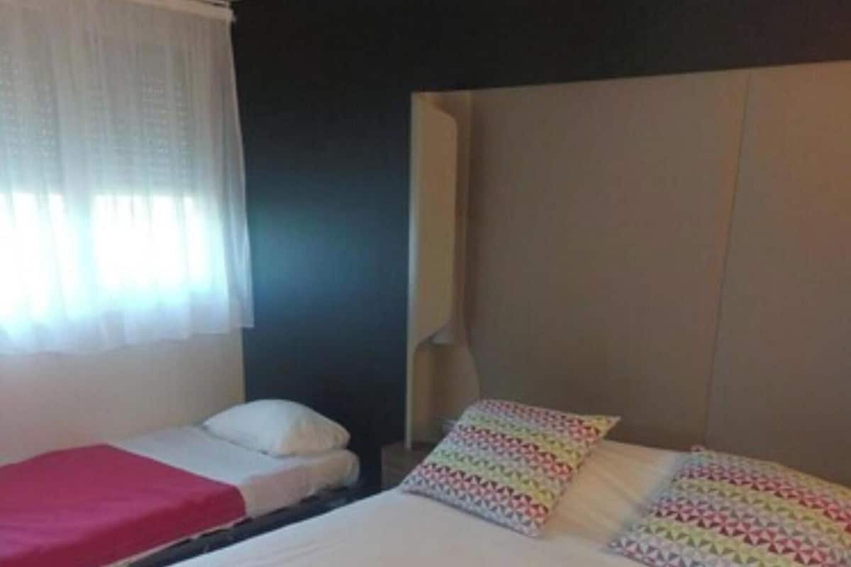 A double bed with white bedding and a red blanket, a bedside table with a lamp, and a window with sheer curtains.