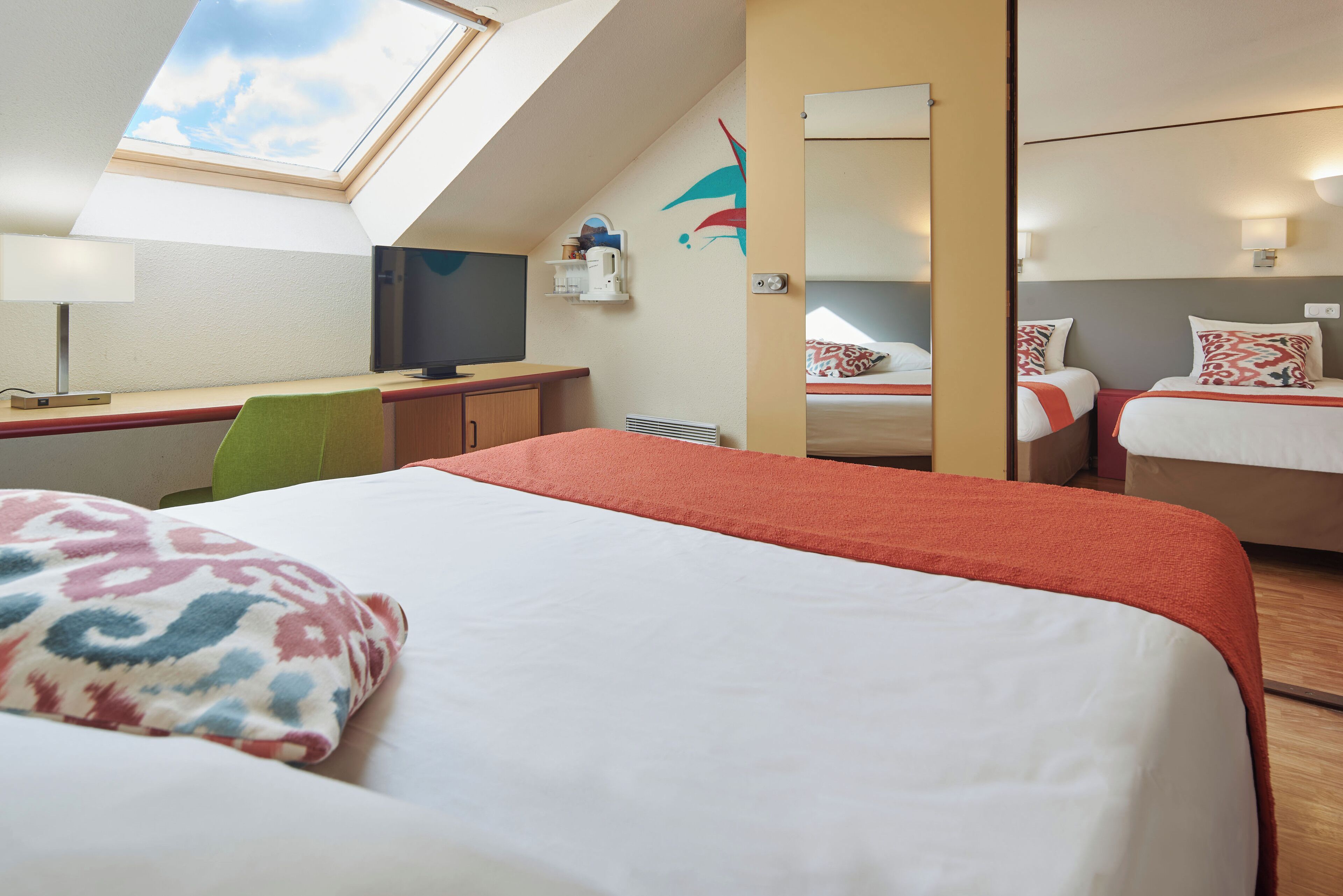 A bedroom with a skylight, a bed with patterned pillows, a desk with a computer, and a mirror.