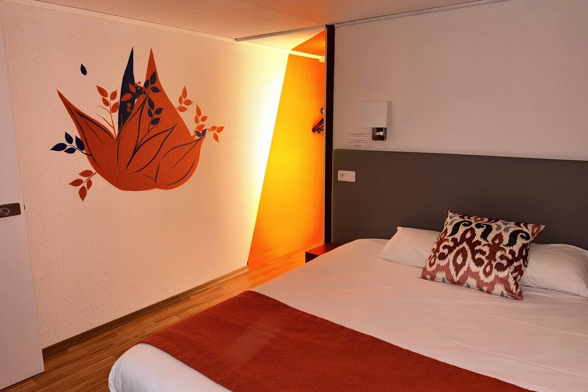 Quarto de hotel com uma cama, um tapete vermelho, uma parede com desenho de folha e uma mesinha de cabeceira.
